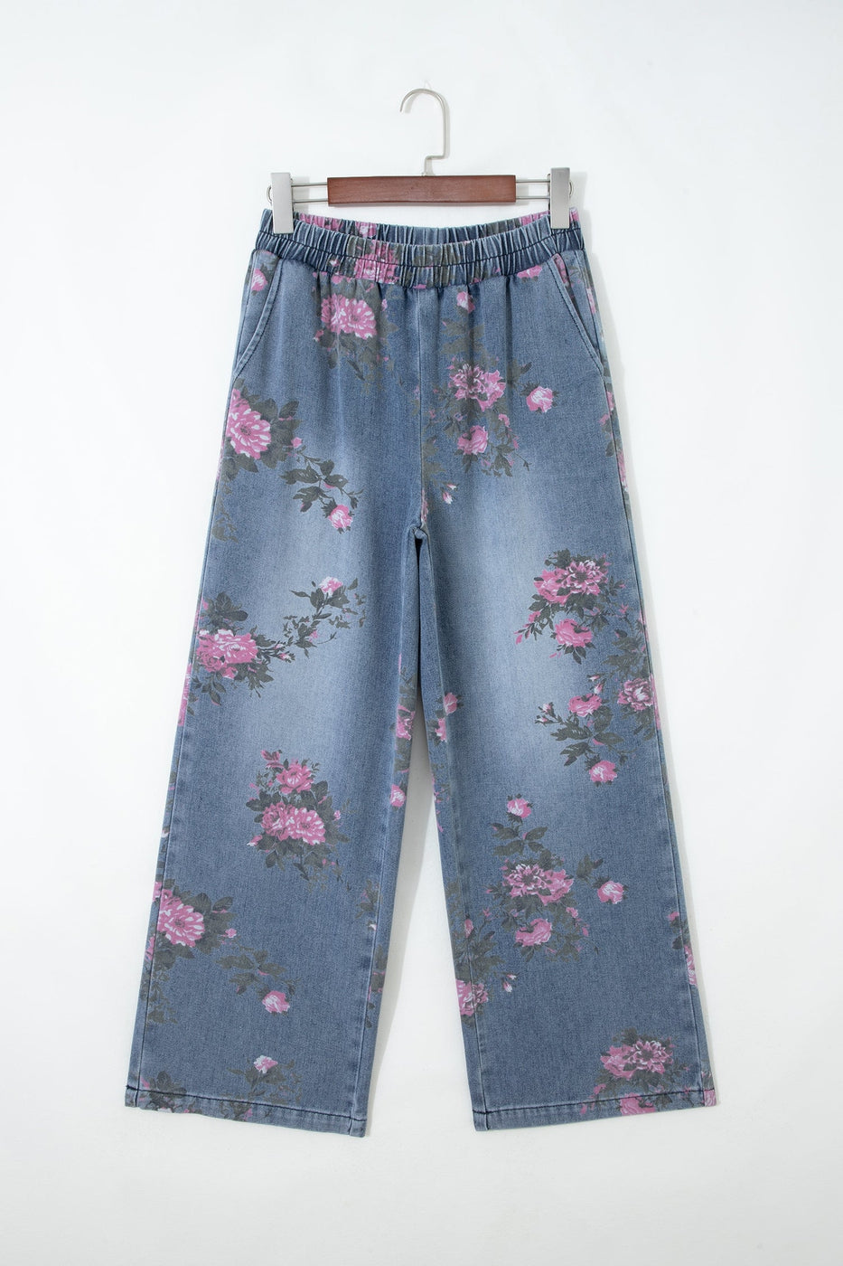 The Floral Pull-On Jeans feature a wide-leg silhouette, elastic waistband, faded blue wash, and pink floral print denim, shown hanging on a wooden hanger against a plain white background.