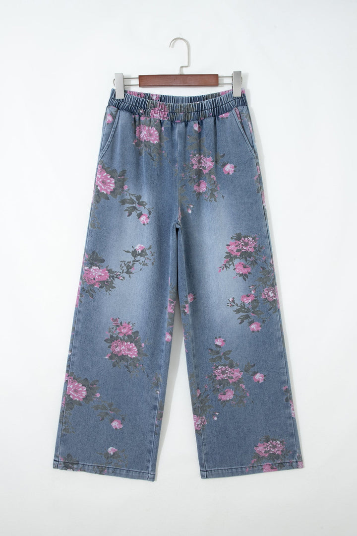 The Floral Pull-On Jeans feature a wide-leg silhouette, elastic waistband, faded blue wash, and pink floral print denim, shown hanging on a wooden hanger against a plain white background.