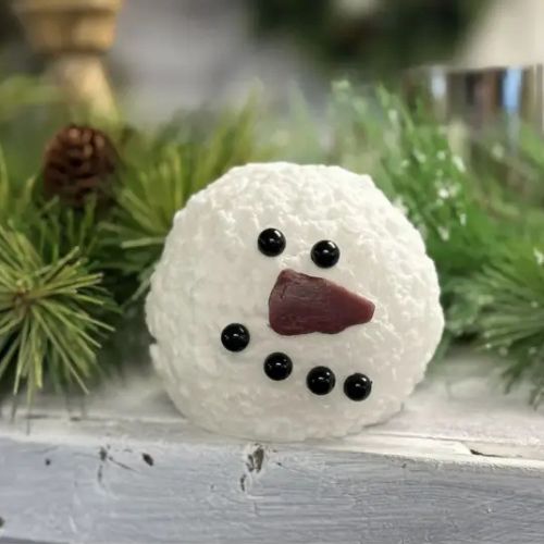 The Snowman Moving Flame Candle, featuring a textured white surface, black bead eyes and mouth, and a red nose, brings festive charm to your holiday décor when displayed among green pine branches on a wooden surface.