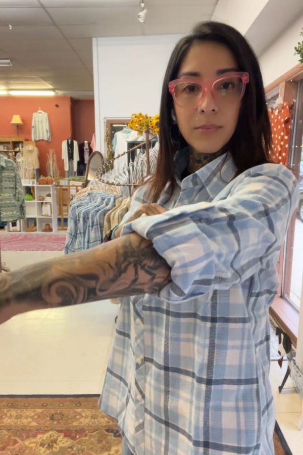 A woman with long dark hair and tattoos rolls up her sleeve in a store, wearing the Plaid Button Shirt and pink glasses. Clothing racks and a cozy boutique setting are visible in the background.