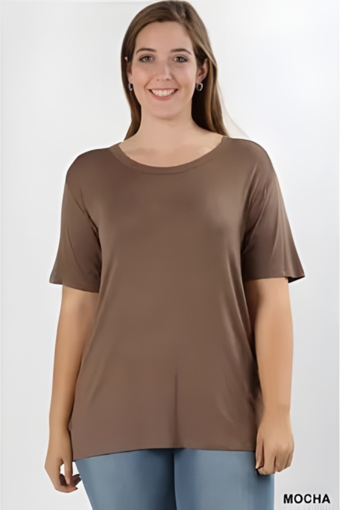 A smiling woman with long brown hair wears a loose-fitting, short-sleeve mocha brown Plus Size Rayon Tee and light blue pants against a plain white background. "MOCHA" appears in the lower right corner.