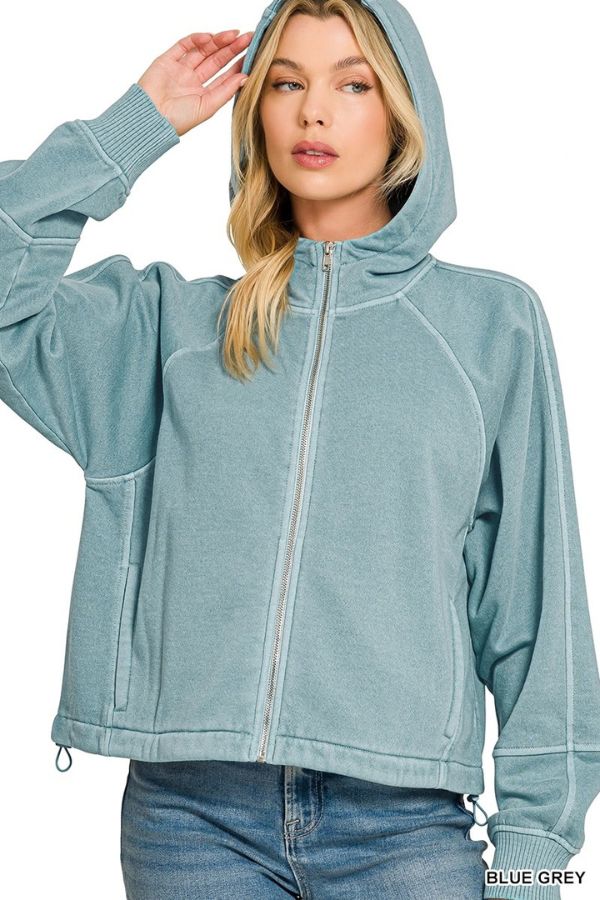 Woman wearing a Pigment Dye Zip Up Hoodie in blue-grey with a relaxed fit, long sleeves, and hood up, touching the edge of the hood while looking slightly to the side.