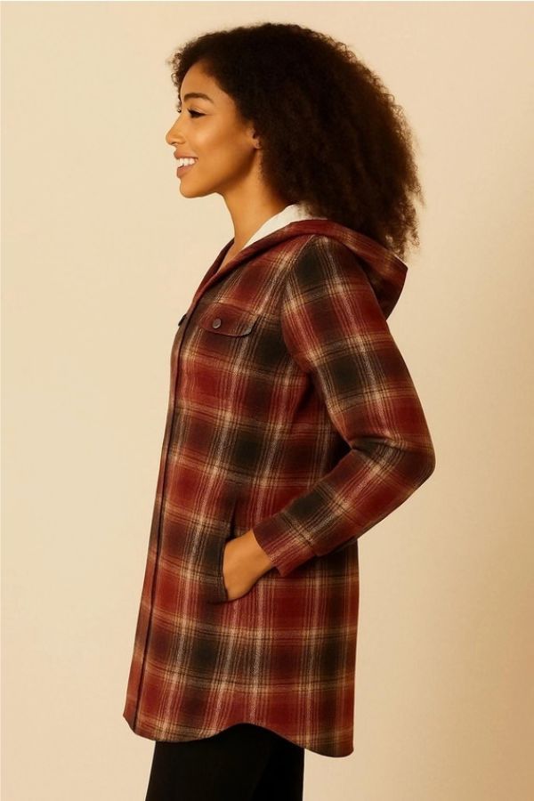 A woman stands sideways, smiling with her left hand in the pocket of a Fur Lined Plaid Hooded Shacket in red, black, and beige tones. This cozy outerwear showcases subtle details that make it ideal for chilly days. Neutral background.
