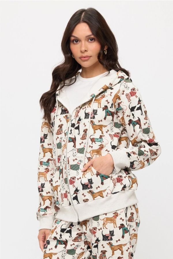 A woman wears the Holiday Dogs Zip Up Hoodie, an oversized zip-up with a vibrant dog print, paired with matching sweatpants. She stands against a plain white background with one hand in her pocket and a neutral expression—a perfect look for dog lovers.