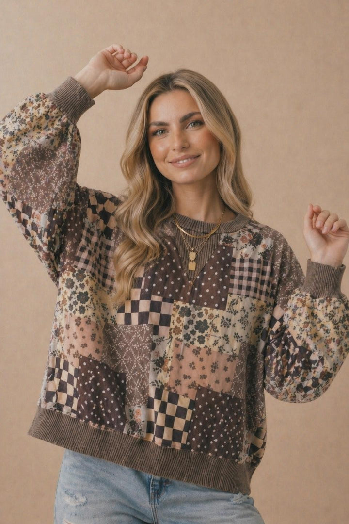 A smiling woman with long, wavy blonde hair poses with both arms raised against a beige background, wearing the Plus Patchwork Pullover—an oversized sweatshirt featuring floral and checkered patterns—paired with light blue jeans and layered gold necklaces.