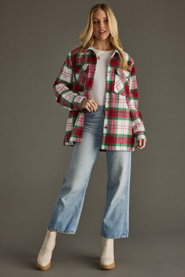 A woman stands against a plain gray background, wearing the Holiday Plaid Cozy Shacket—a red and green plaid, oversized jacket with a button-front closure—layered over a white top, light blue jeans, and cream ankle boots.