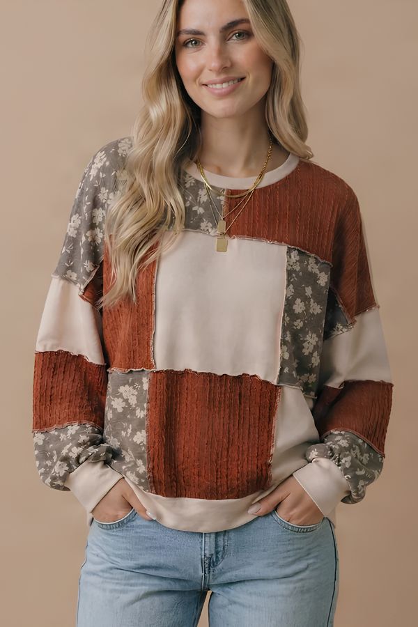 A smiling woman wears the Color Block Knit Top, featuring a relaxed fit and patchwork design in earthy tones with fall floral print patches. She pairs it with light blue jeans and layered gold necklaces, standing against a beige background.