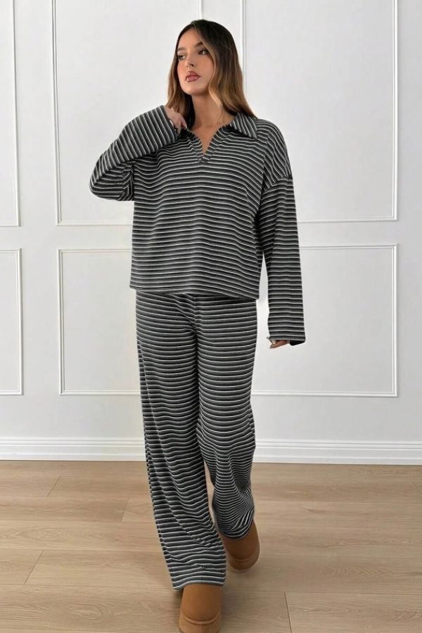 A person stands indoors posing against a white paneled wall and light wood floor, wearing the Striped Collared Lounge Set—a loose black and white striped two-piece featuring a collared pullover top and matching wide-leg pants—paired with brown slippers.