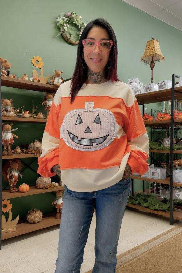 A woman with glasses and dyed hair wears a Gingham Pumpkin Sweatshirt and jeans, standing in a store decorated with autumn and pumpkin ornaments.