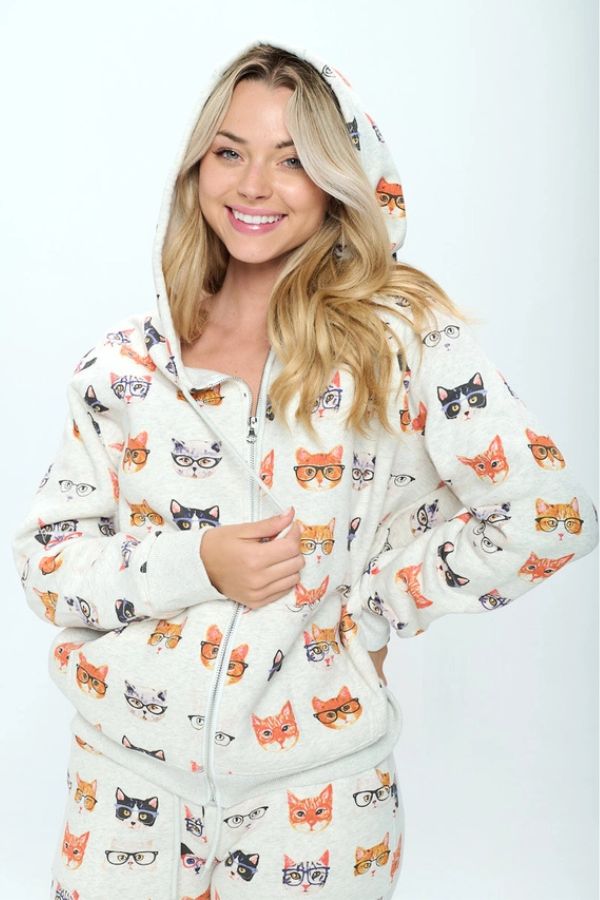 A smiling person wearing an oversized, cream-colored Cats in Glasses Zip Hoodie featuring a playful print of cats wearing eyeglasses.