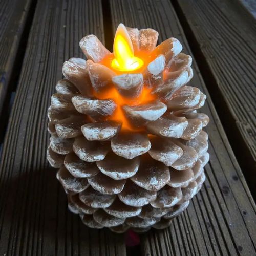 A Pine Cone Moving Flame Candle with a sculpted pinecone design sits on a wooden surface, its realistic moving flame casting a warm yellow-orange glow that highlights the candle’s textured details.