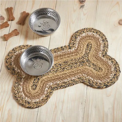 Two stainless steel pet bowls rest on the Braided Bone Shaped Rug, which features a durable braided construction and a playful bone-shaped design. The rug is set against a wood floor background, with three dog bone-shaped treats nearby.