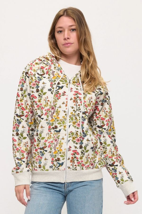A person wearing the Floral Hummingbird Zip Hoodie—a white zip-up adorned with a vibrant floral print—paired with blue jeans and a white shirt stands against a plain light background. The relaxed fit of the hoodie enhances the laid-back, stylish vibe.