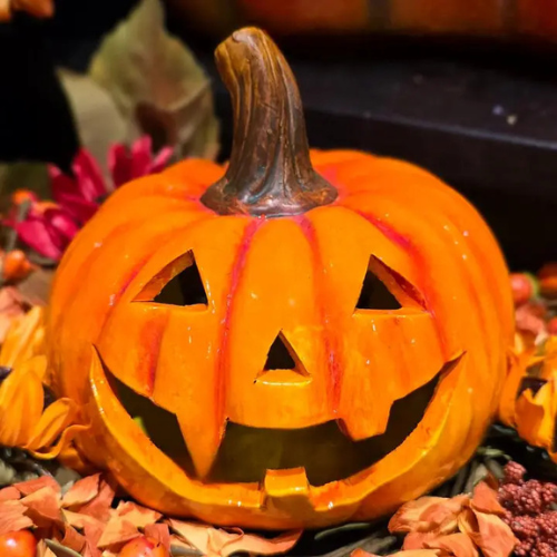 The Harvest Harry Jack-O’-Lantern, featuring a carved smiling face, sits among autumn leaves and flowers, making it a charming piece of fall decor.