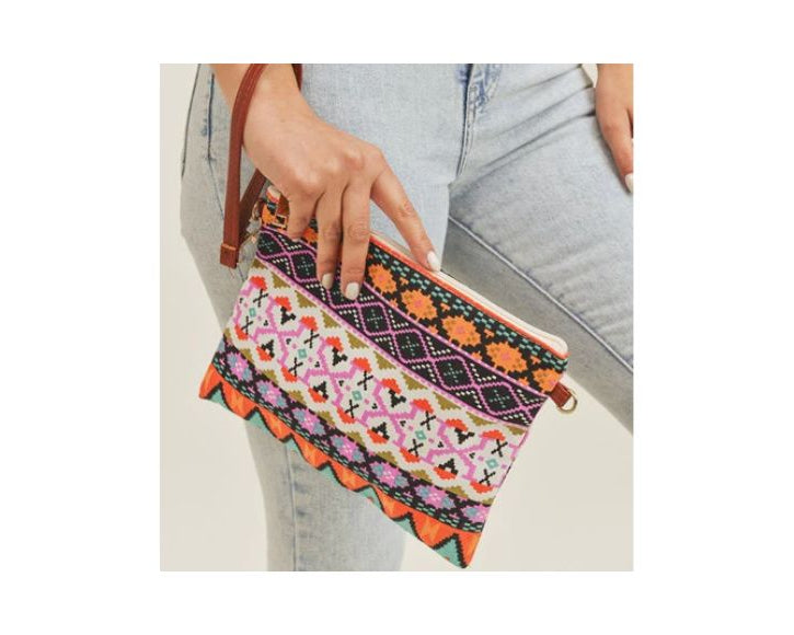 A woman wearing light-wash jeans holds a vibrant, rectangular clutch bag featuring a colorful Aztec-inspired geometric pattern in shades of pink, orange, black, and white, with a brown wrist strap.