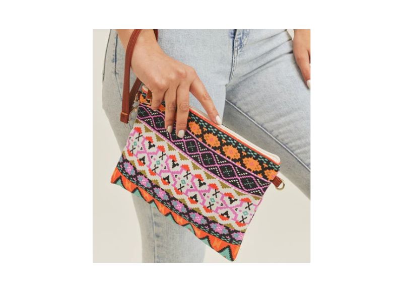 A woman wearing light-wash jeans holds a vibrant, rectangular clutch bag featuring a colorful Aztec-inspired geometric pattern in shades of pink, orange, black, and white, with a brown wrist strap.