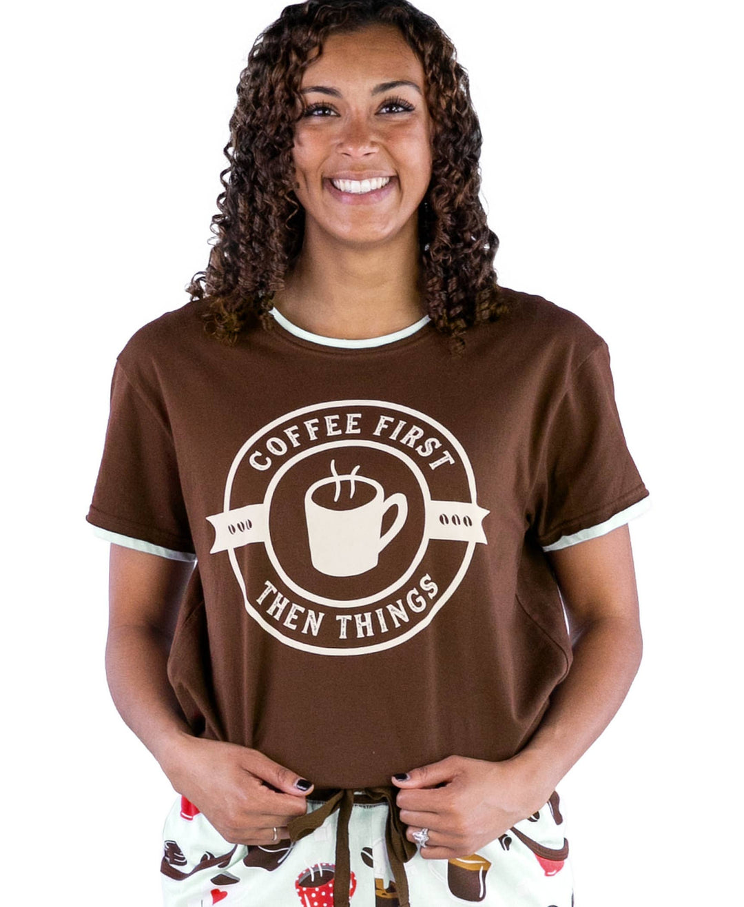 A smiling woman with curly hair wears a Dog Mom, Cat Nap & More Pajama Top—a comfy brown cotton tee featuring a coffee cup graphic and the words "Coffee First Then Things"—while standing against a white background.