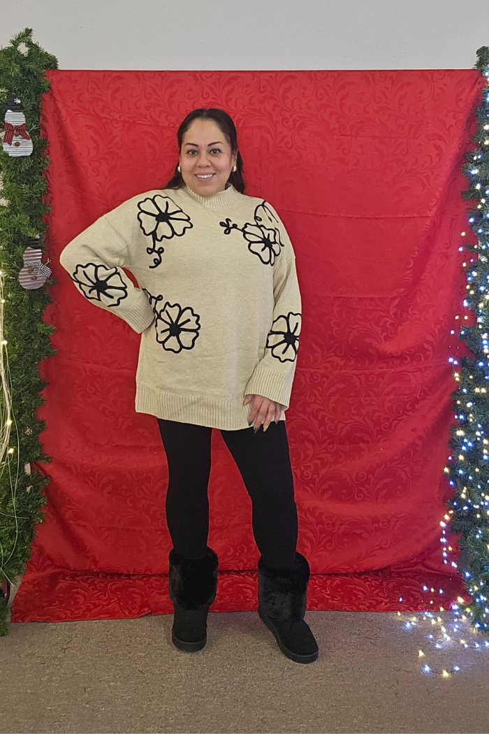 A woman smiles before a red patterned backdrop with holiday greenery and lights, wearing the Floral Embroidered Knit Sweater paired with black leggings and boots—ideal for chic boutique winter style.