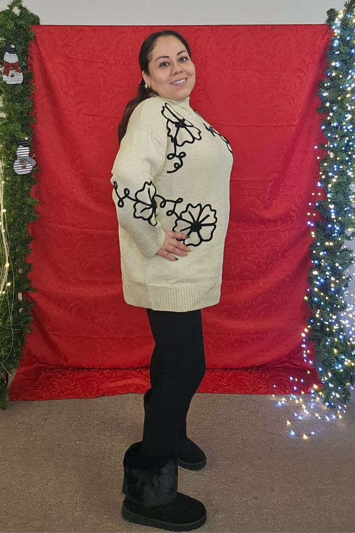 A woman smiles in front of a red backdrop, wearing the Floral Embroidered Knit Sweater with black leggings and boots. She poses sideways amid decorated, lit greenery.