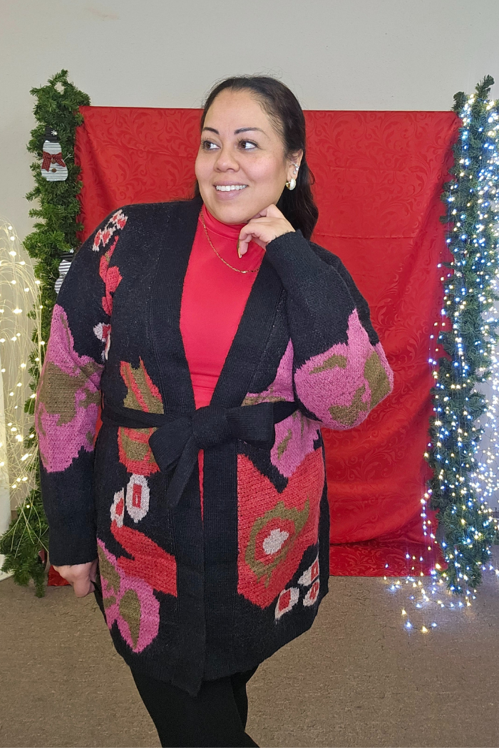 A woman smiles and poses in front of a festive red backdrop, wearing a red top paired with the Floral Belted Knit Cardigan featuring bold pink, red, and green patterns.