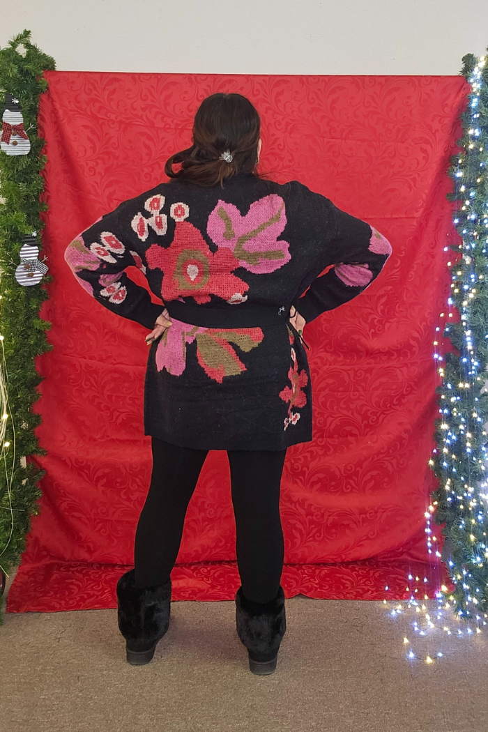 A person faces away from the camera before a red patterned backdrop, wearing the Floral Belted Knit Cardigan with bold pink and red designs, black leggings, and fur boots. Greenery and string lights frame the scene.