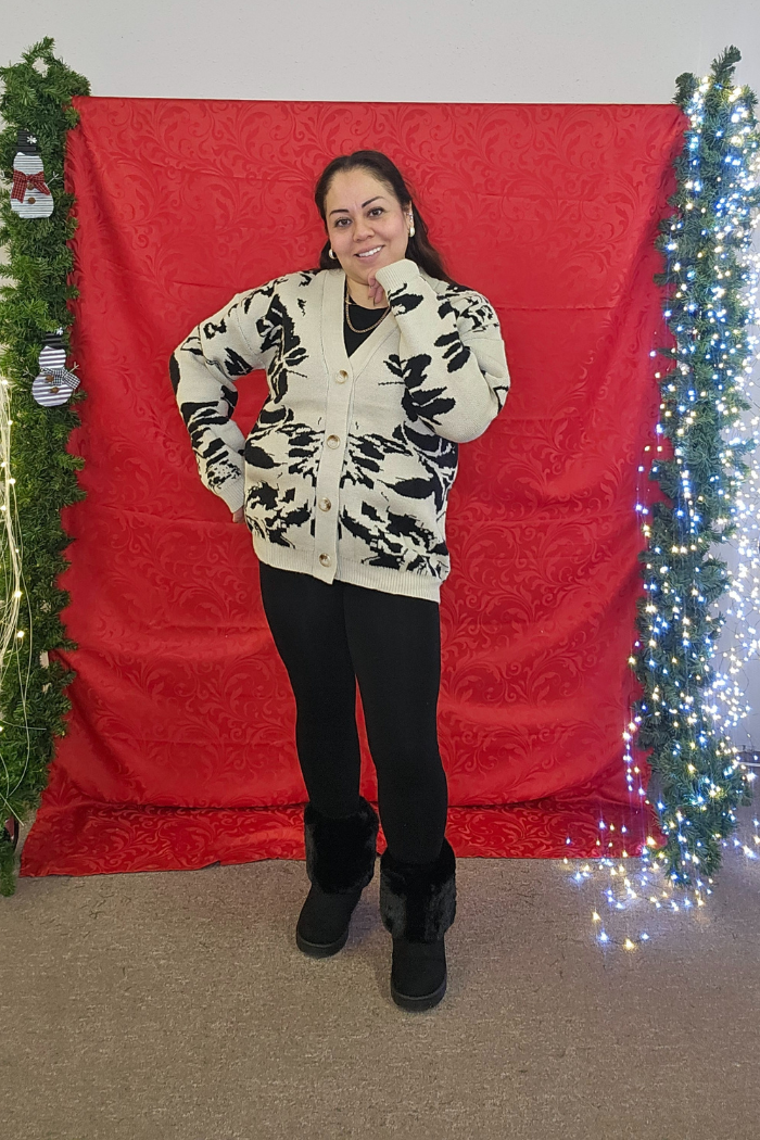A woman poses in front of a red backdrop wearing the Multi Print Button Cardigan, black leggings, and black fur boots. She stands amid vertical garlands adorned with holiday decorations and string lights.