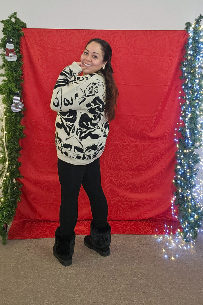 A woman wearing the Multi Print Button Cardigan, black leggings, and black boots smiles in front of a red backdrop adorned with green garlands and white string lights.