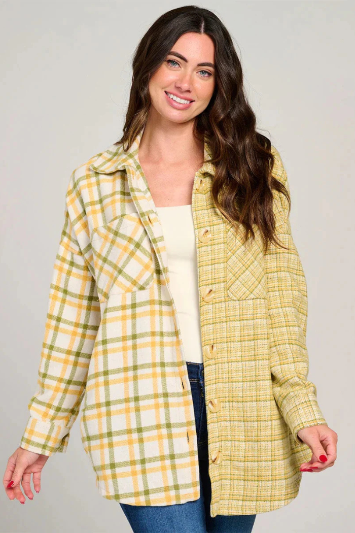 A smiling woman with long brown hair wears a two-toned plaid shacket featuring a yellow, white, and green plaid pattern on the left side and a finer yellow and white plaid pattern on the right side. She is also wearing a white top and blue jeans, standing against a plain light background.