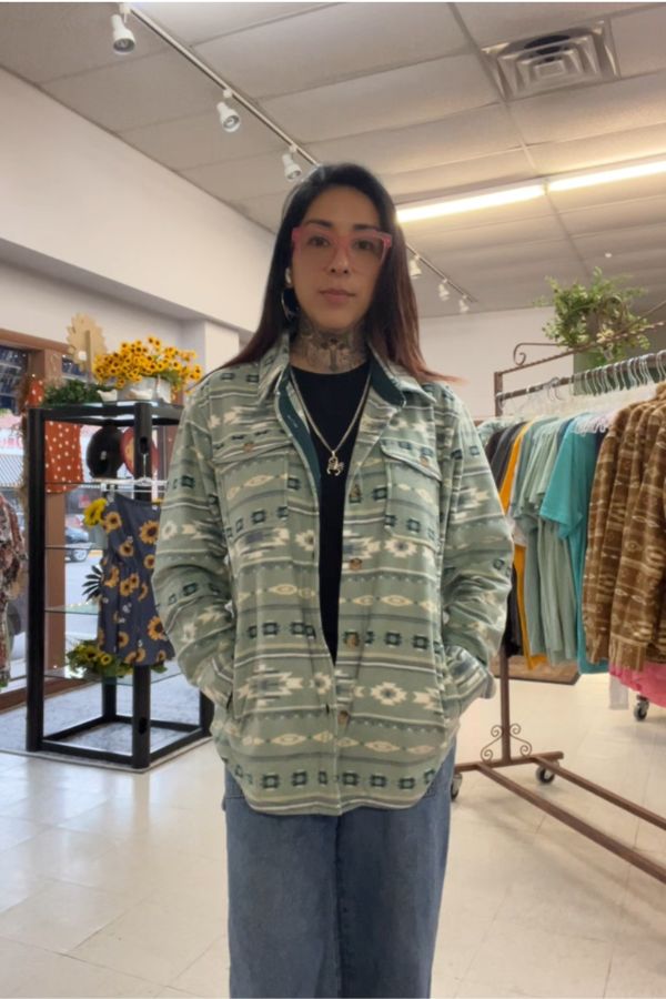 A person with long dark hair and glasses stands indoors in a clothing store, hands in pockets, wearing the Southwestern Fleece Shacket featuring a light green geometric pattern. Clothing racks and sunflowers are visible in the background.