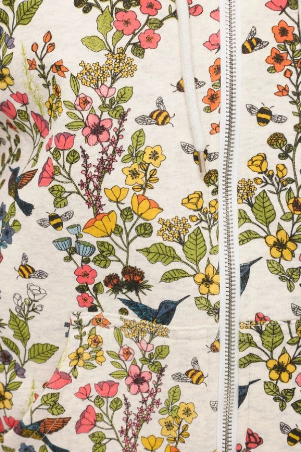Close-up of the Floral Hummingbird Zip Hoodie, made from a light-colored fabric adorned with vibrant yellow, pink, and orange flowers, as well as bees, butterflies, and blue birds. The relaxed fit hoodie features a vertically running white zipper.
