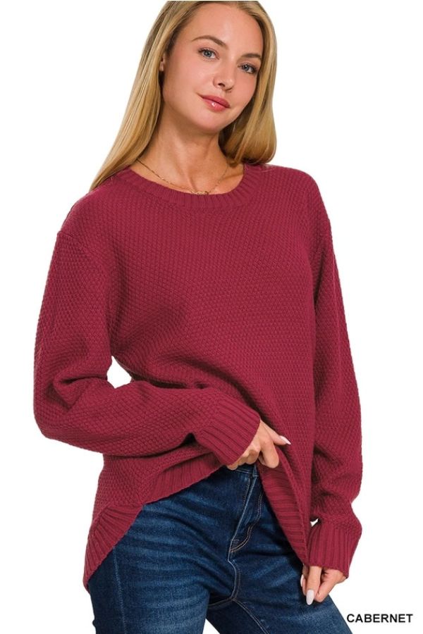A woman with long blonde hair wears the Everyday Crew Neck Sweater in dark red, paired with blue jeans. She stands with one hand in her pocket and the other holding the hem of her sweater, looking slightly to the right and smiling softly.