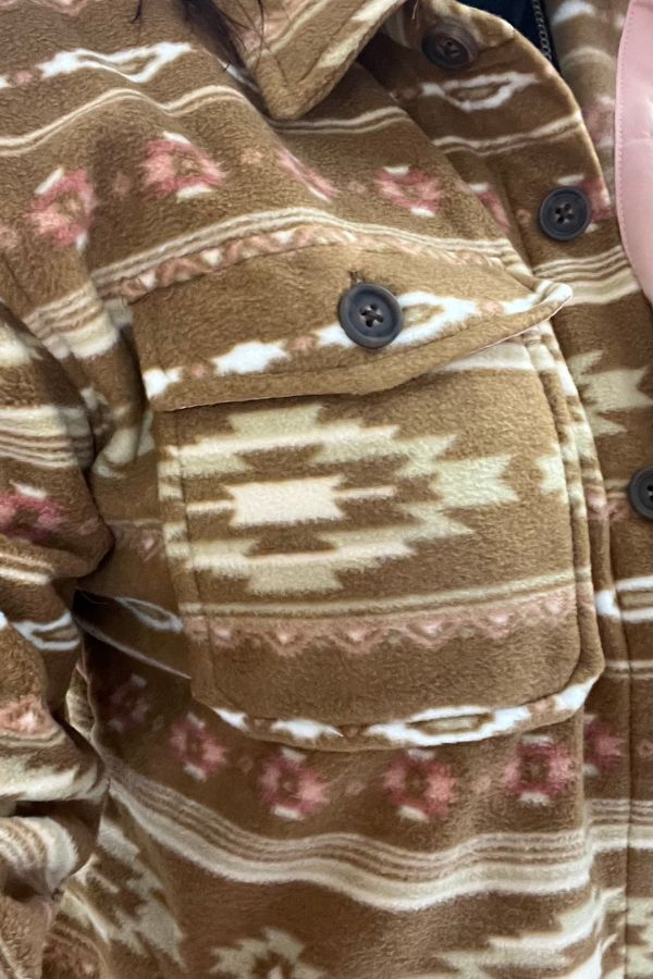 Close-up of the Southwestern Fleece Shacket in brown, showcasing a pocket and geometric pattern in cream and pink tones. This cozy shacket features dark buttons and a visible collar, adding a stylish touch to its design.
