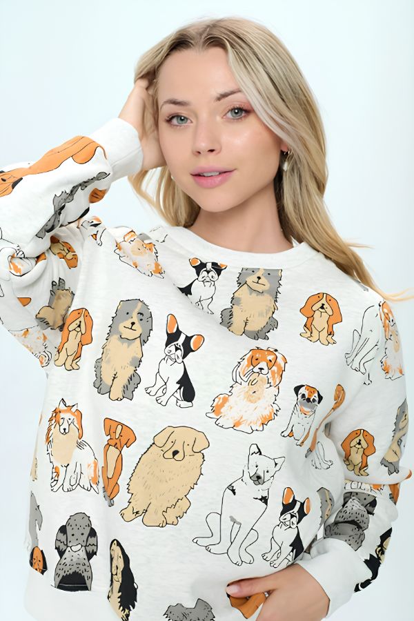 A woman with long blonde hair models the Dog Print Crewneck Sweatshirt, which features illustrations of various dog breeds in orange, brown, and black tones—perfect for dog lovers—against a plain light background.