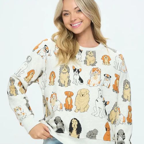 A woman with long blonde hair smiles while wearing the Dog Print Crewneck Sweatshirt decorated with playful cartoon dog illustrations, posing against a simple light background—an ideal gift for any dog lover.