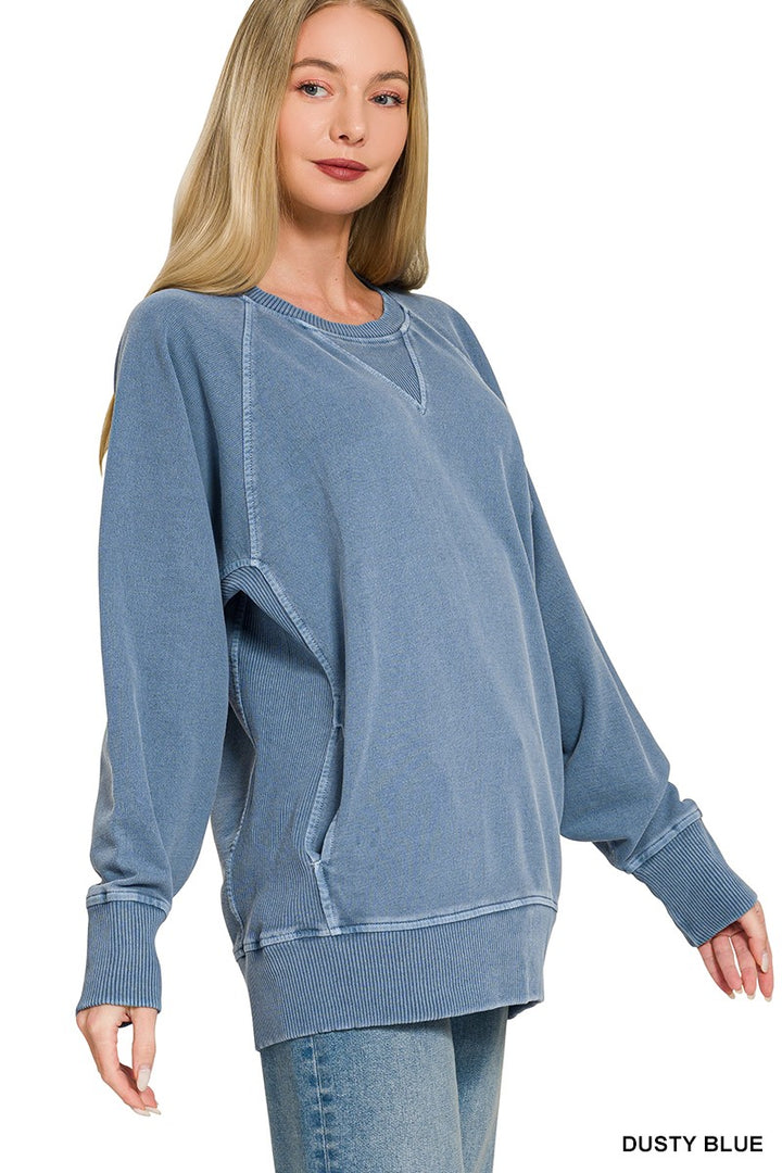 A woman with long blonde hair wears a relaxed fit Pigment Dyed Pullover in dusty blue and blue jeans, standing against a white background.