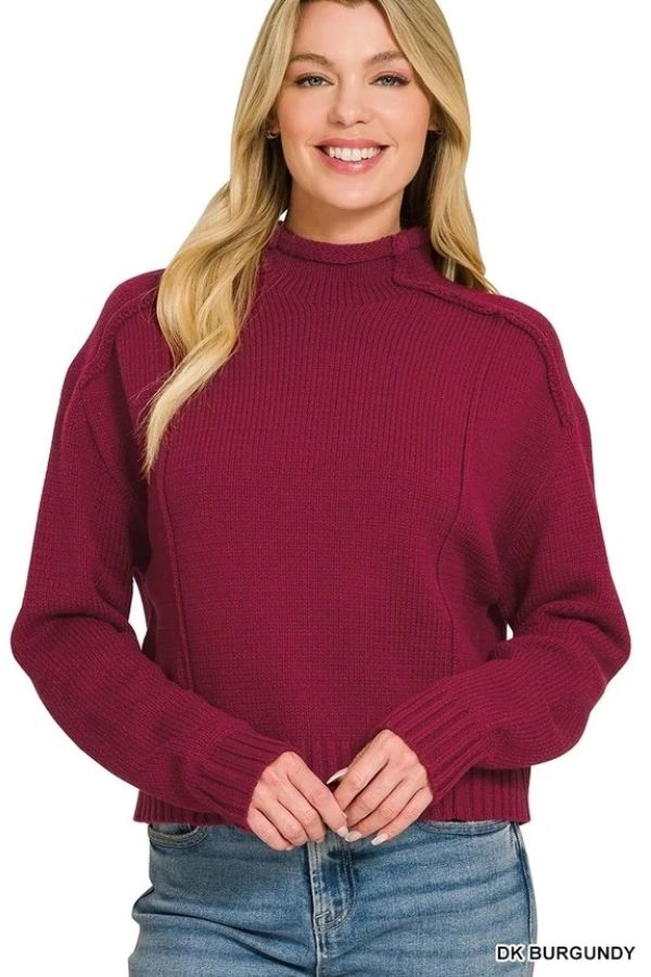 A smiling woman wearing a burgundy Mock Neck Exposed Seam Sweater, a relaxed-fit knit with a mock neckline and visible vertical seams on the shoulders and chest.