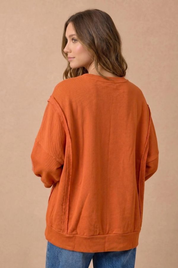 A woman with wavy brown hair, wearing the Plaid Thanks Patchwork Sweatshirt and blue jeans, stands facing away but looks over her shoulder against a plain beige background.