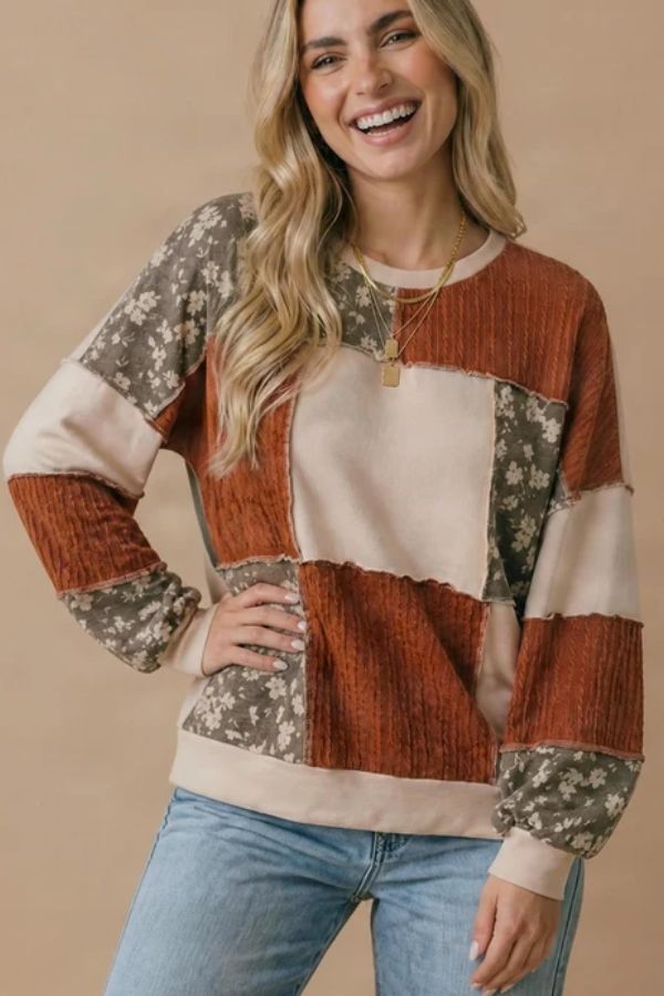 Woman smiling, wearing a relaxed fit Color Block Knit Top featuring rust, beige, and floral patchwork-style patterns, paired with light blue jeans, standing in front of a neutral beige background.