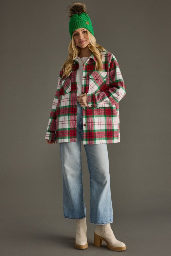 A woman stands against a plain gray background, wearing the Holiday Plaid Cozy Shacket—a red and green plaid oversized jacket with a button-front closure—styled with a green knit hat featuring a pom-pom, white top, light blue jeans, and beige heeled boots for an effortlessly cozy fall look.
