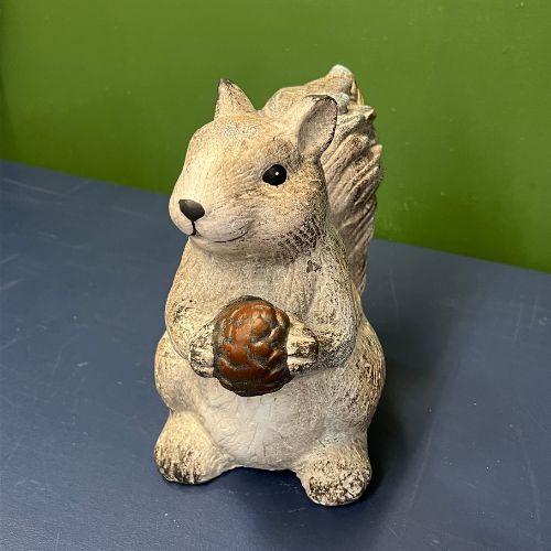 The Scamper Squirrel Figurine, crafted from ceramic and depicted holding a brown pinecone, stands on a dark blue surface against a green wall backdrop—making it an ideal addition to farmhouse décor or fall decorating.