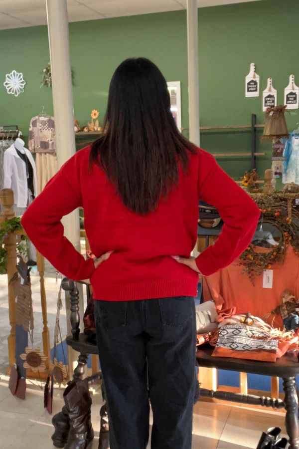 A person with long dark hair, wearing the Snowman Christmas Knit Sweater in festive red and black pants, stands indoors with their hands on their hips, facing away from the camera. The background features shelves, clothing, and decorative items.