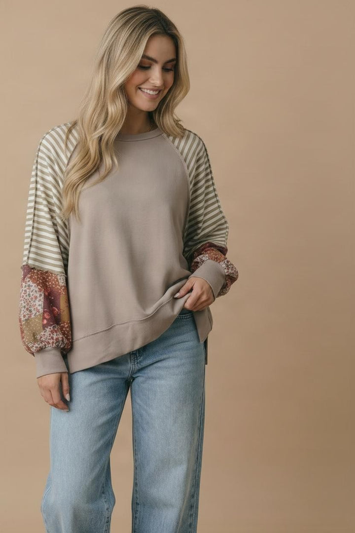 A woman with long blonde hair wears the Plus Patchwork Sleeve Top, featuring taupe French terry and mixed print sleeves, paired with light blue jeans. She stands against a beige background, smiling and looking down.