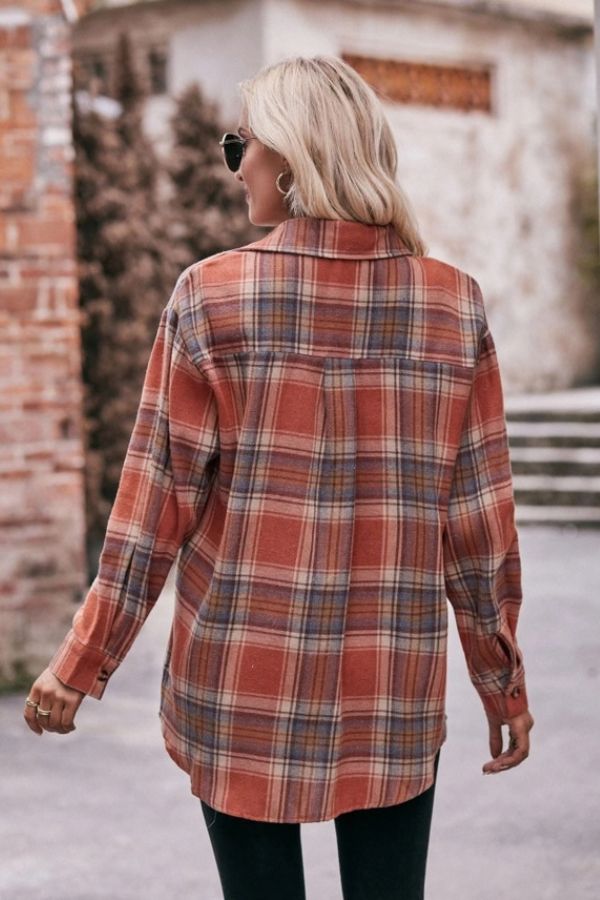 A person with long blonde hair, wearing sunglasses and an Oversized Plaid Button Shirt in orange tones, stands outdoors with their back to the camera. Brick walls and greenery are visible in the background.