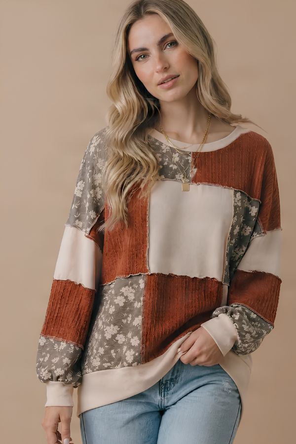 A woman models the Color Block Knit Top, featuring fall-inspired floral and patchwork sections in rust and cream. She pairs it with light blue jeans and a gold necklace. Her long, wavy blonde hair cascades over her shoulders as she stands against a beige background.