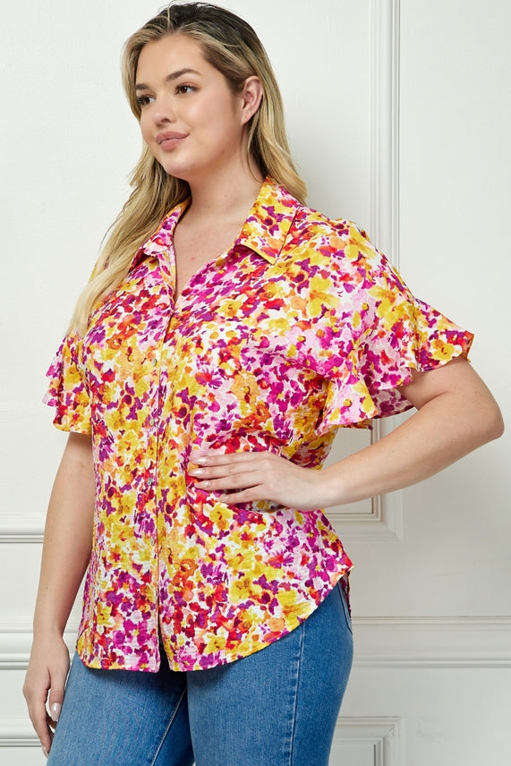 A woman with long blonde hair wears the Plus Floral Button Blouse and blue jeans, standing against a white paneled wall.
