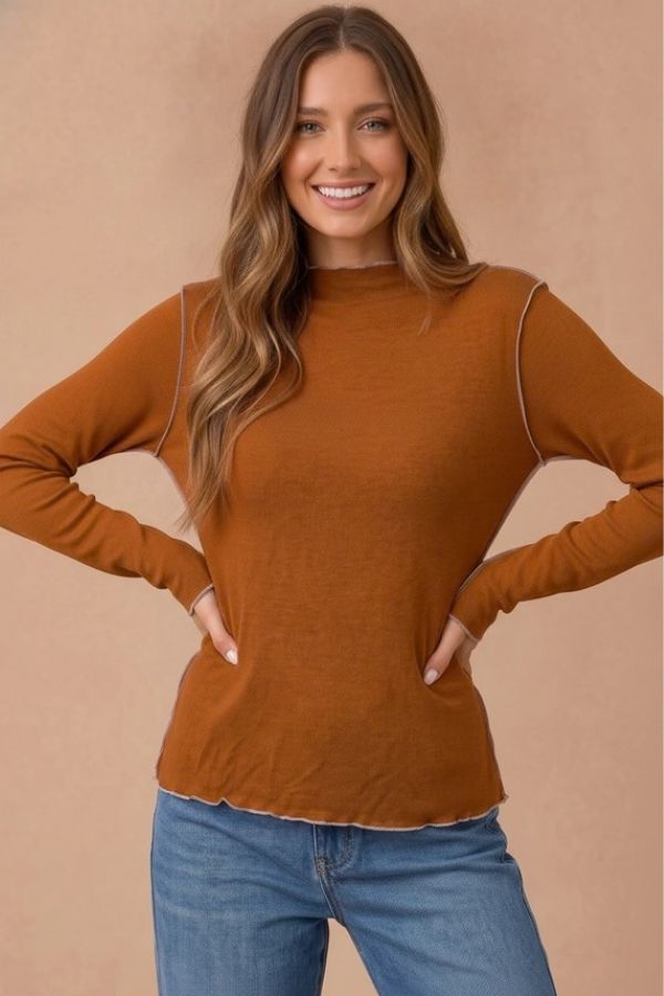 A woman wears a rust-colored Curvy Frilled Mock Neck Hacci Top with a frilled mock neckline, long sleeves, exposed light-colored seam detailing, and blue jeans.
