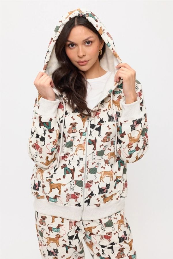 Wearing the Holiday Dogs Zip Up Hoodie and matching pants, featuring a vibrant all-over dog print, she stands against a plain white background, looking at the camera and holding the strings of her hoodie—an ideal outfit for dog lovers.