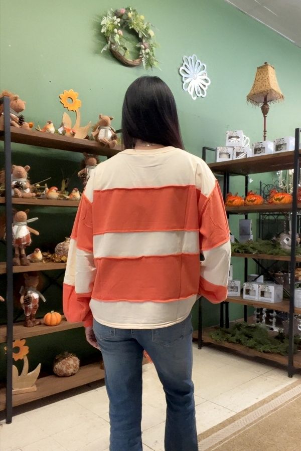 Wearing the Gingham Pumpkin Sweatshirt, along with jeans and a red-and-white striped shirt, a person with long dark hair stands facing away in a store decorated with autumn-themed items and shelves lined with plush toys.