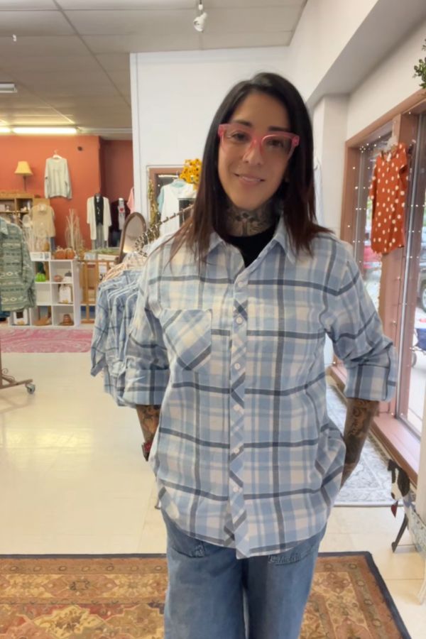 A person with long dark hair, pink glasses, and tattoos is wearing the Plaid Button Shirt with jeans, standing in a boutique filled with clothing and home décor.
