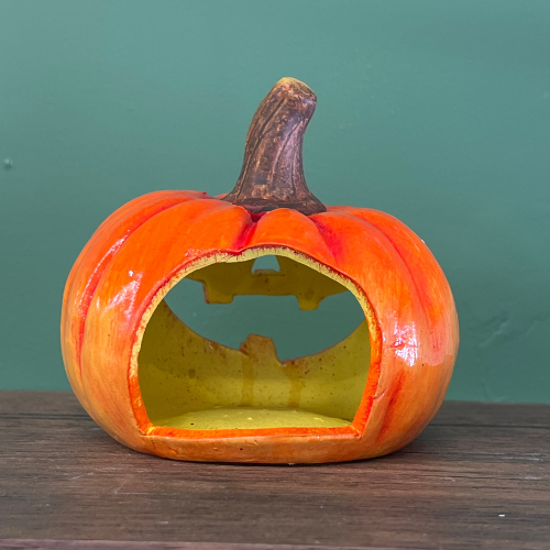 The Harvest Harry Jack-O’-Lantern sits on a wooden surface against a green wall, its orange ceramic exterior featuring a carved-out mouth, brown stem, and yellow interior—perfect as charming fall decor.
